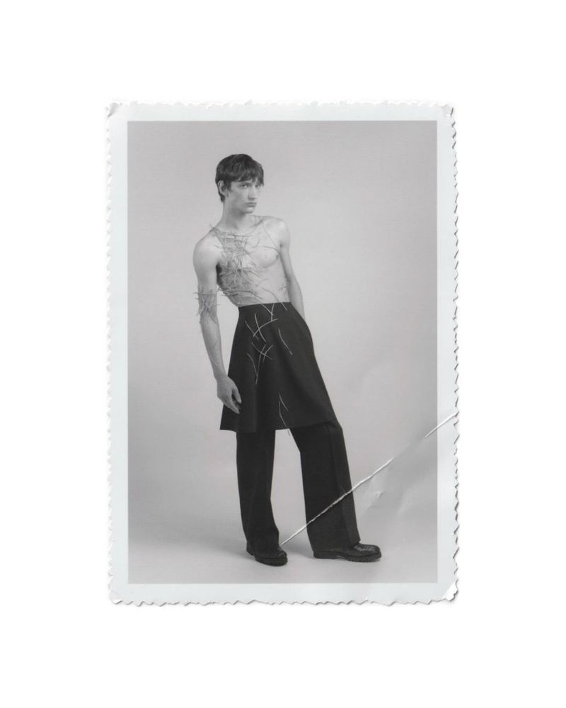 Monochromatic fashion photo of a slender male model in avant-garde clothing posing in a white studio, emphasizing silhouette and fabric.