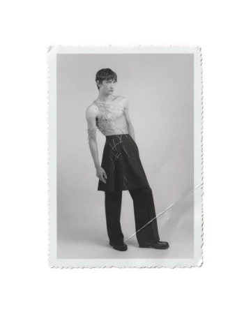 Male model wearing a black skirt over trousers with thread detailing against a seamless grey backdrop in a vintage border print.