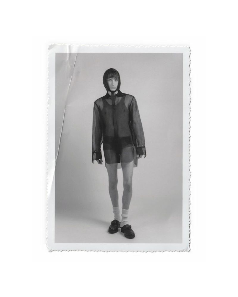 Black and white portrait of a slender adult male model in a hooded sheer shirt, shorts, socks, and shoes, posing in a photo studio.