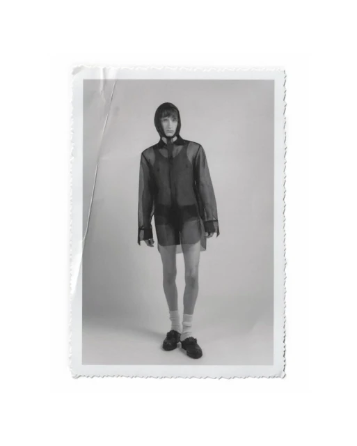 Black and white portrait of a slender adult male model in a hooded sheer shirt, shorts, socks, and shoes, posing in a photo studio.