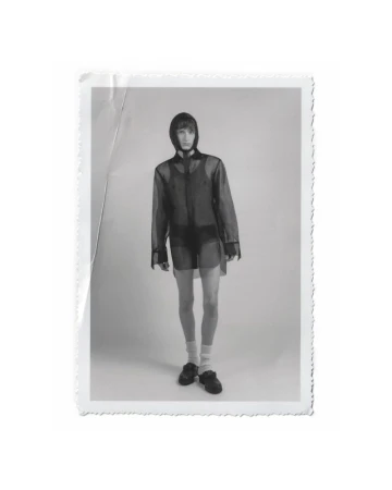 Black and white portrait of a slender adult male model in a hooded sheer shirt, shorts, socks, and shoes, posing in a photo studio.