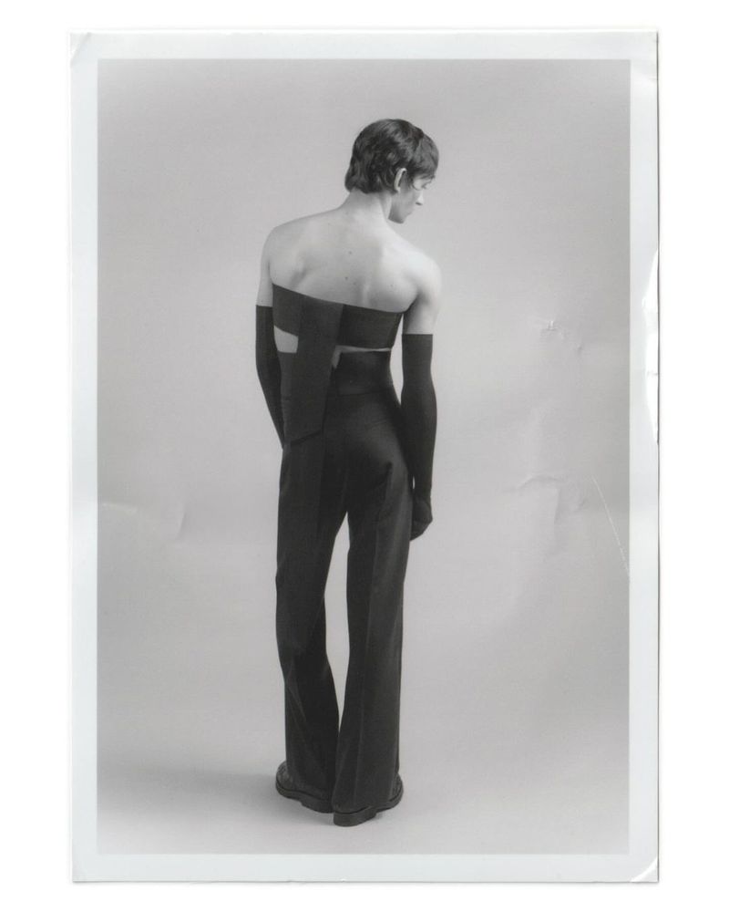 Back view of a slender adult male model in a black avant-garde two-piece outfit with arm coverings, standing in a photo studio setting.