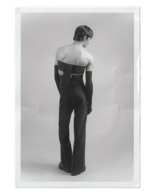 Back view of a slender adult male model in a black avant-garde two-piece outfit with arm coverings, standing in a photo studio setting.