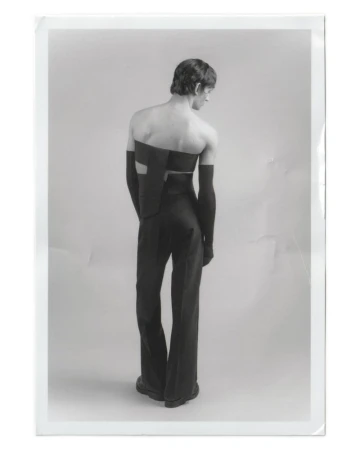 Back view of a slender adult male model in a black avant-garde two-piece outfit with arm coverings, standing in a photo studio setting.