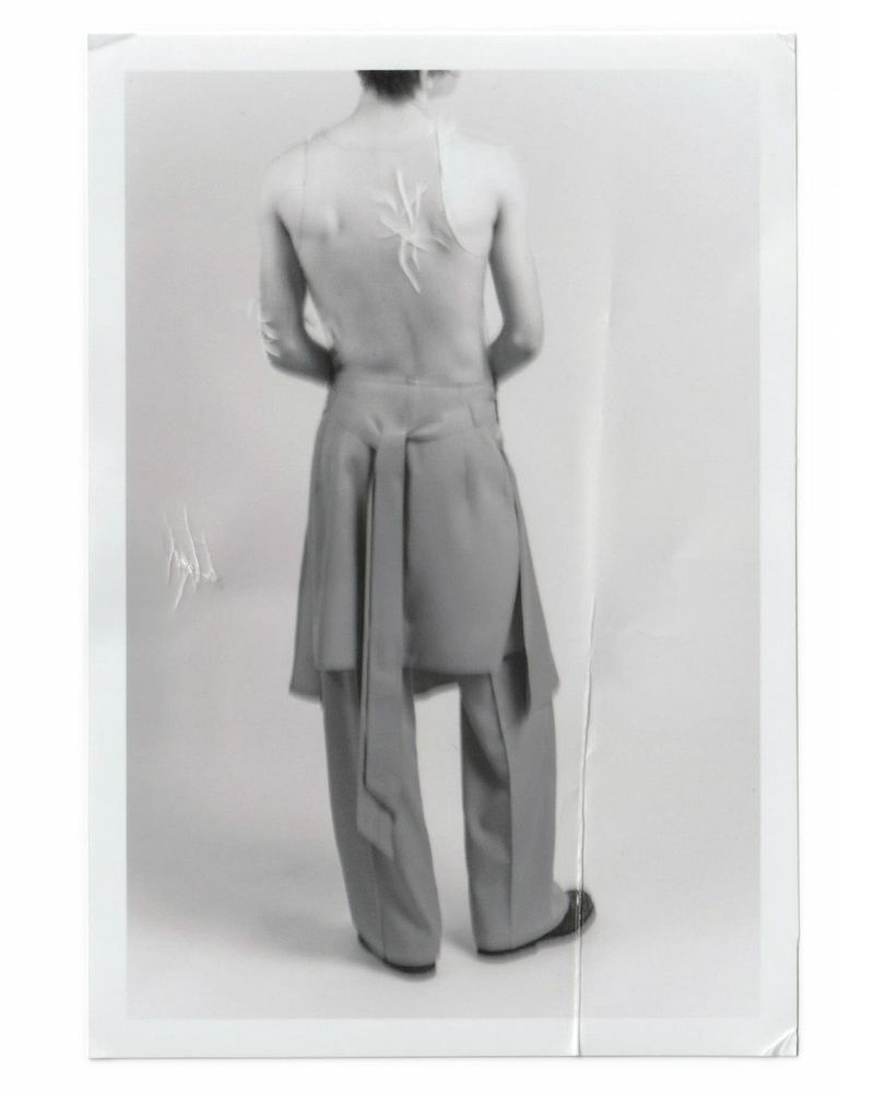 An adult fashion model with a spider design on their back, wearing grey tailored trousers and black shoes, poses in a photo studio.