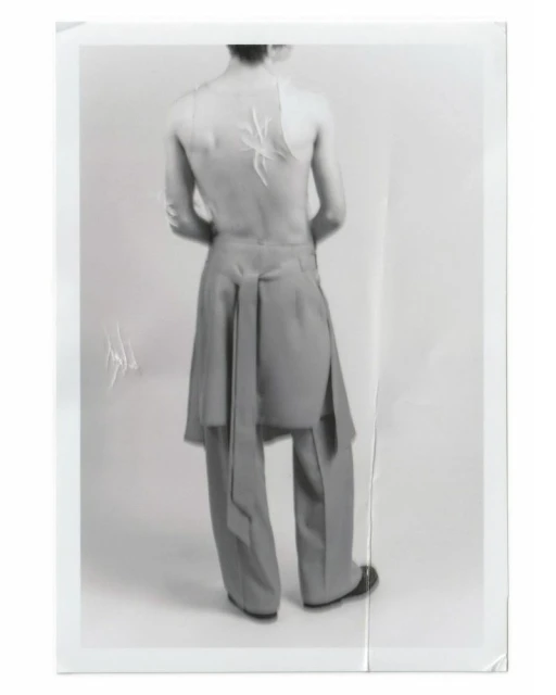 An adult fashion model with a spider design on their back, wearing grey tailored trousers and black shoes, poses in a photo studio.