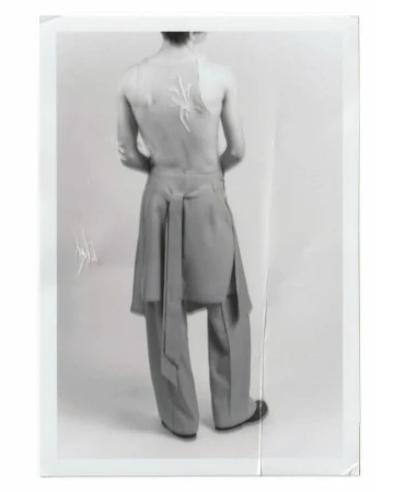 An adult fashion model with a spider design on their back, wearing grey tailored trousers and black shoes, poses in a photo studio.