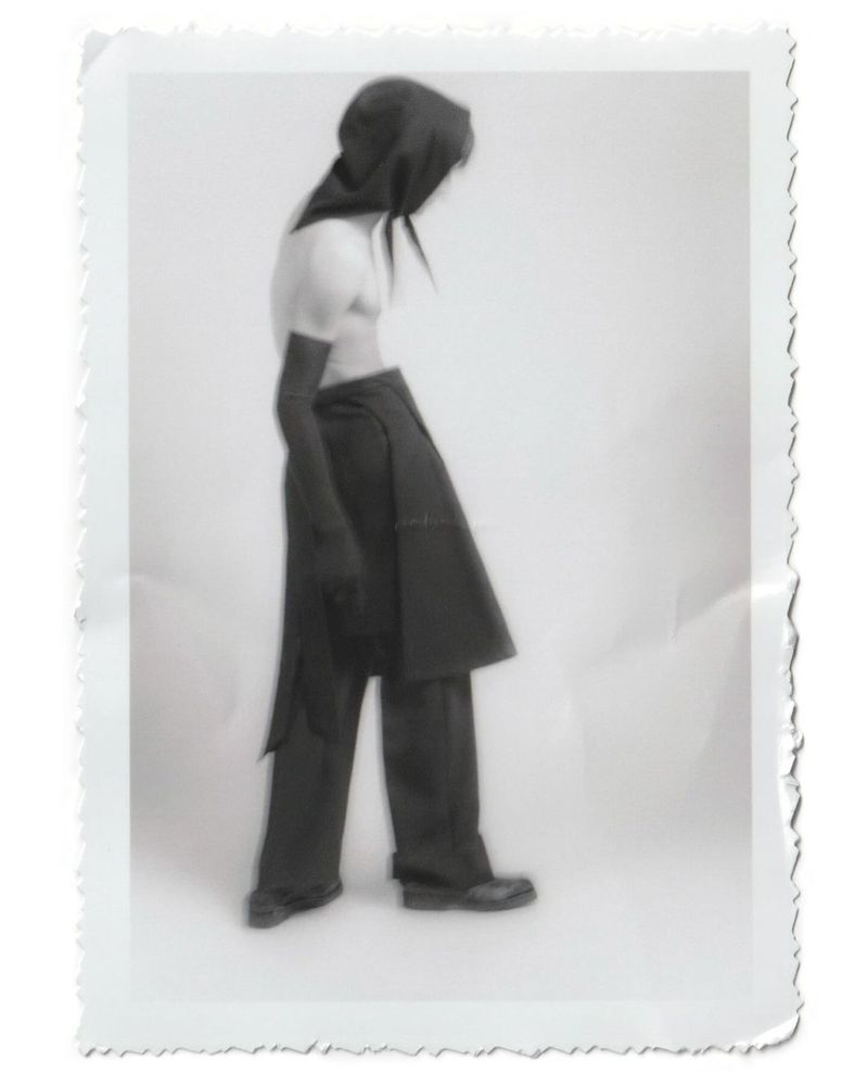 Person wearing a dark hooded outfit in a studio setting, captured in a vintage-style photograph with a white background and textured border.