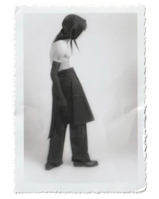 Person wearing a dark hooded outfit in a studio setting, captured in a vintage-style photograph with a white background and textured border.