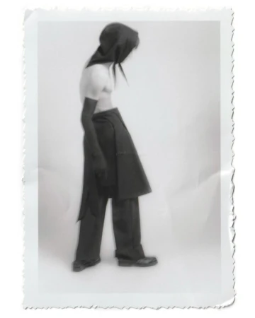 Person wearing a dark hooded outfit in a studio setting, captured in a vintage-style photograph with a white background and textured border.