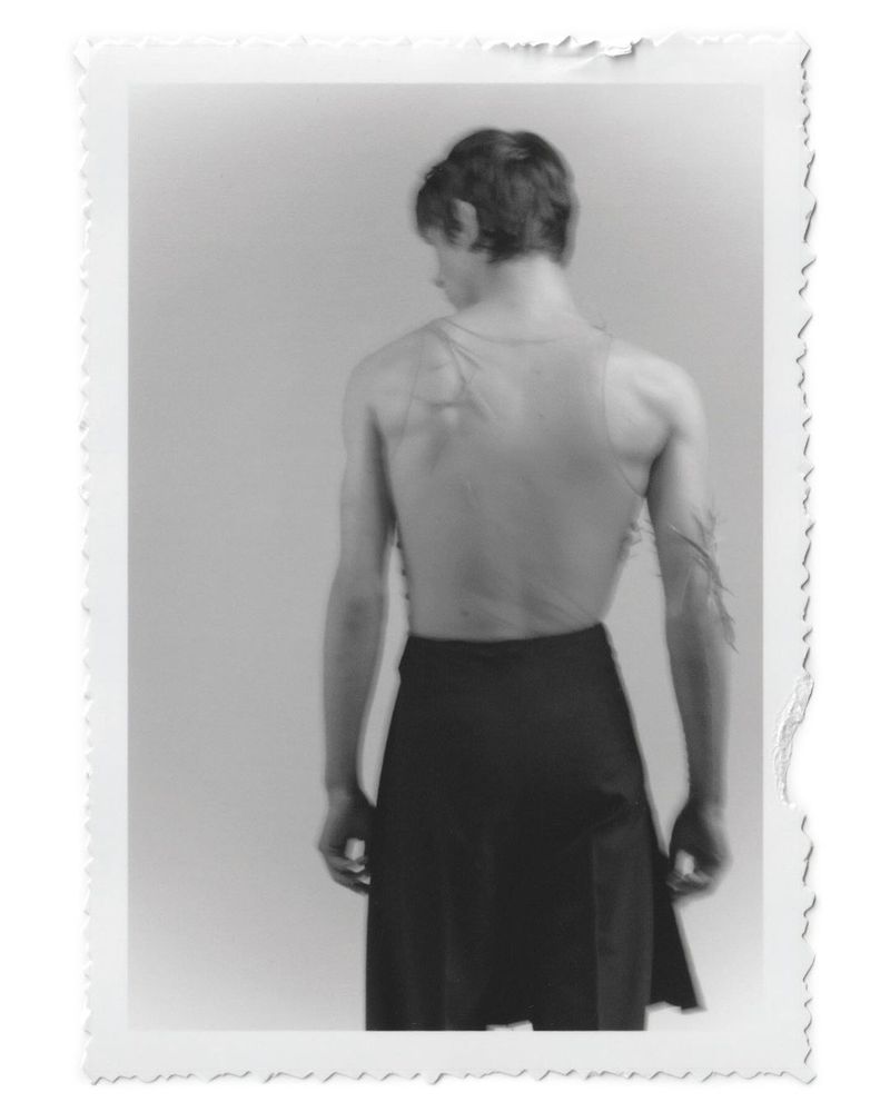 Black and white photograph of a slender, muscular adult male from the rear, wearing a dark skirt, against a plain background.