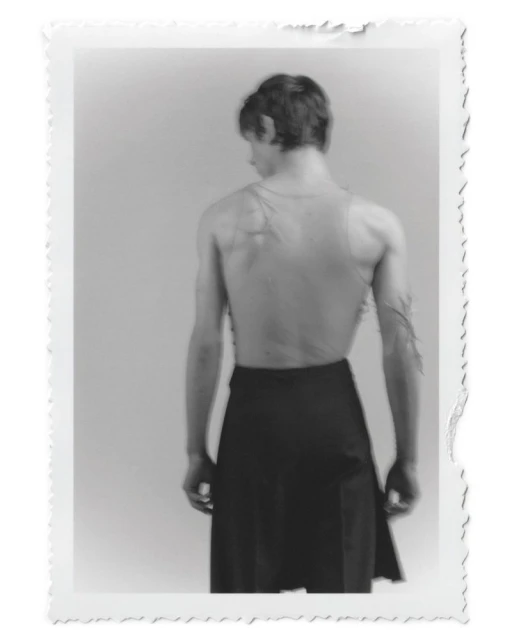 Black and white photograph of a slender, muscular adult male from the rear, wearing a dark skirt, against a plain background.