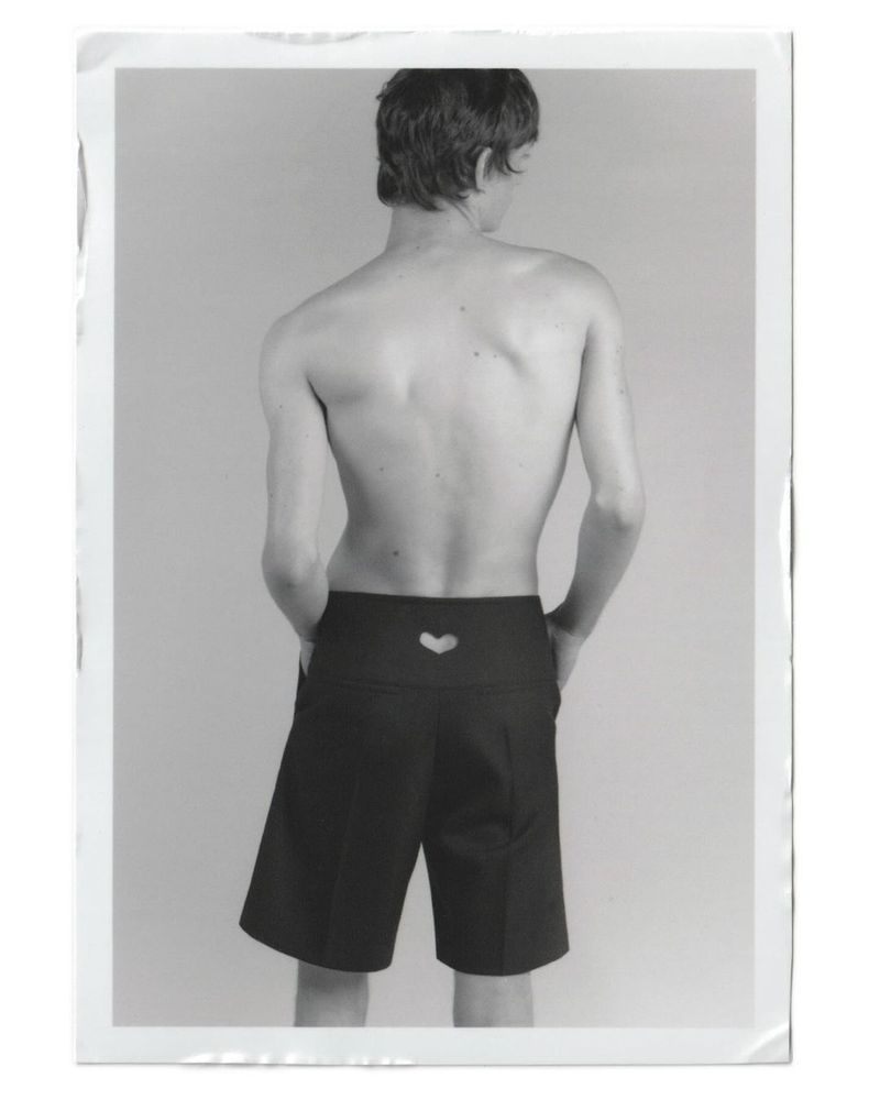 Back view of a slender, topless adult male wearing black shorts with a heart cutout, standing in a brightly lit photo studio.