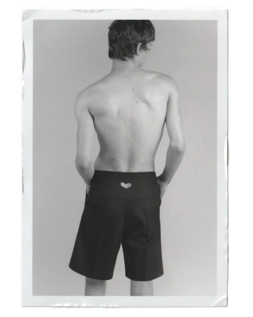 Back view of a slender, topless adult male wearing black shorts with a heart cutout, standing in a brightly lit photo studio.
