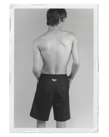 Back view of a slender, topless adult male wearing black shorts with a heart cutout, standing in a brightly lit photo studio.