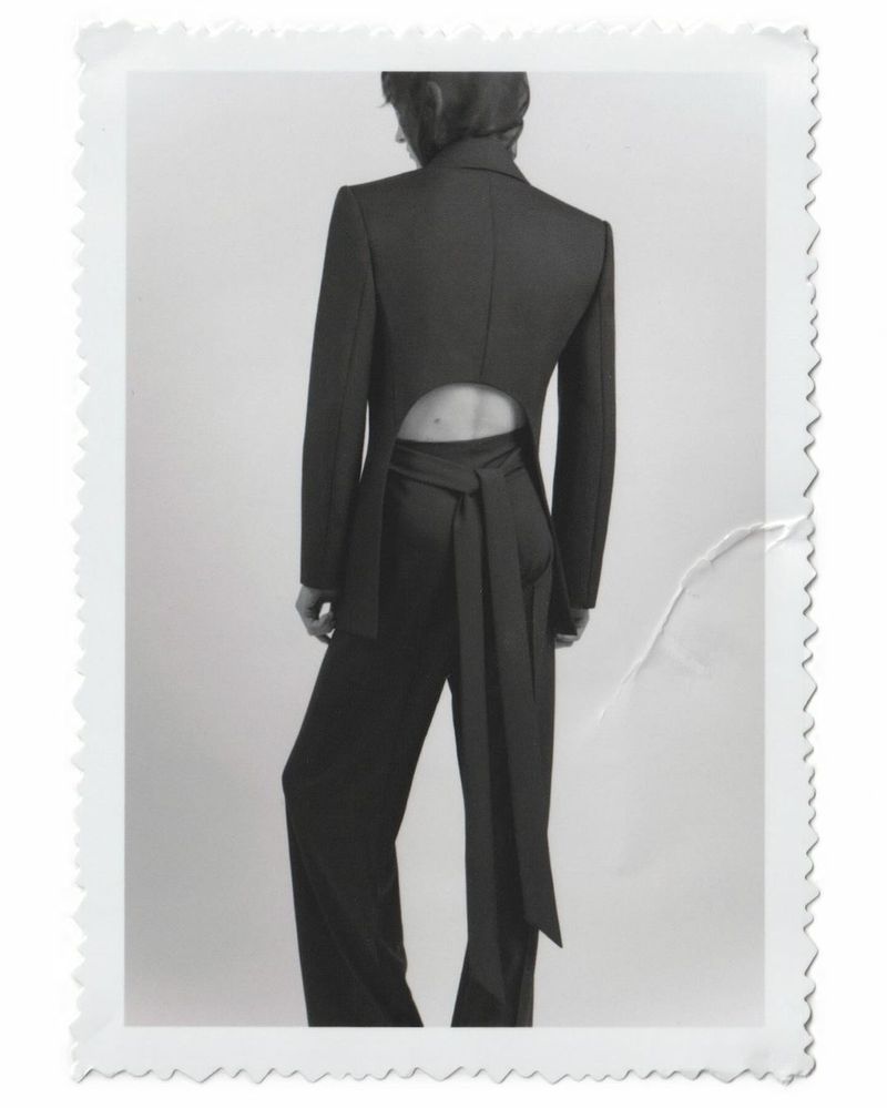 Back view of a woman in a dark pant suit with a tie at the waist, against a white backdrop in a studio setting. Fashion editorial.