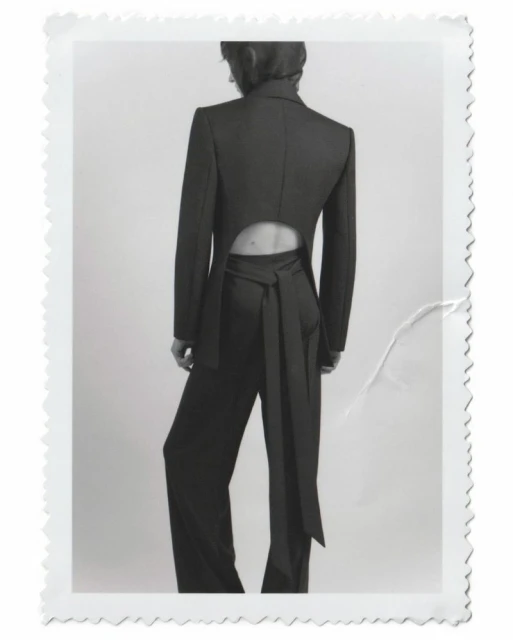 Back view of a woman in a dark pant suit with a tie at the waist, against a white backdrop in a studio setting. Fashion editorial.