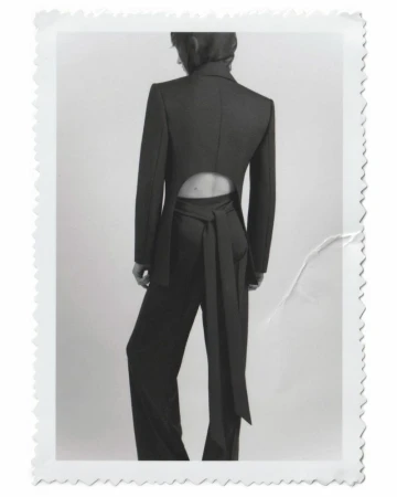 Back view of a woman in a dark pant suit with a tie at the waist, against a white backdrop in a studio setting. Fashion editorial.