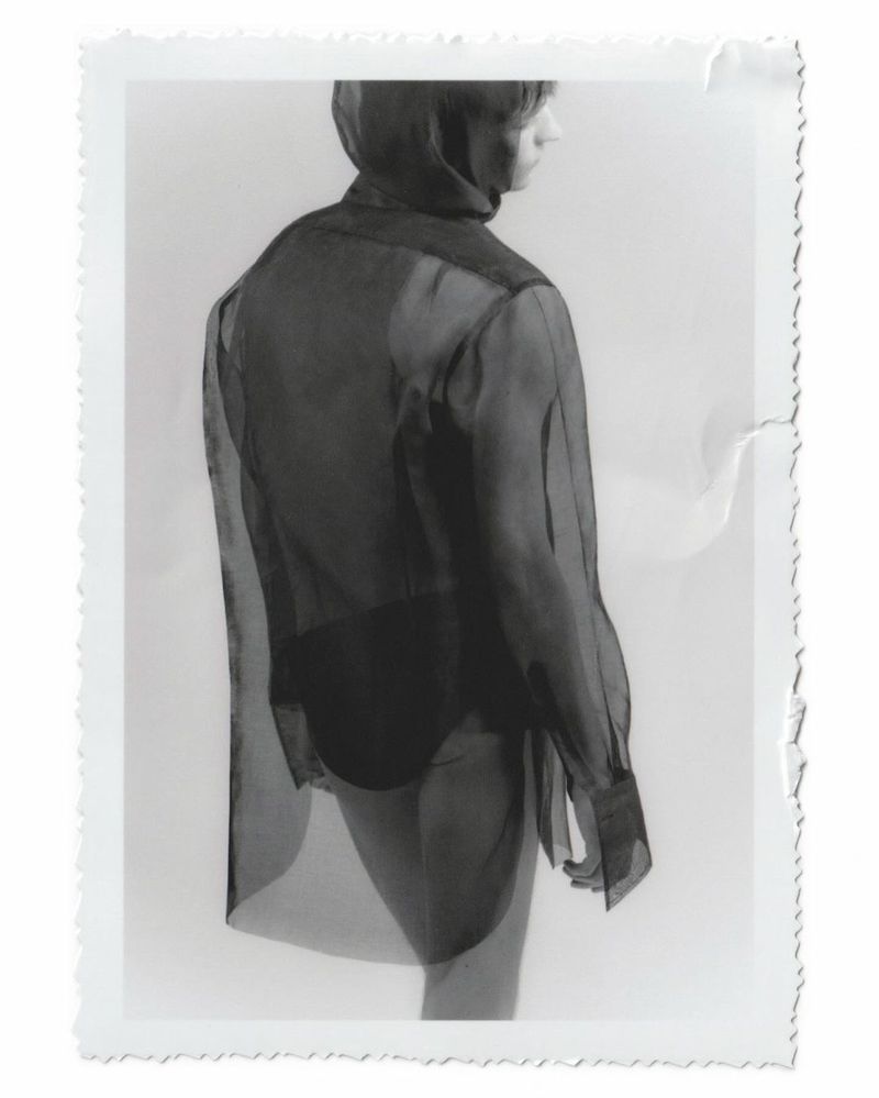 Back view of a slim person wearing a sheer black blouse and dark bottoms, captured in a studio setting under bright, neutral lighting.