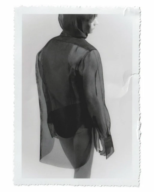 Back view of a slim person wearing a sheer black blouse and dark bottoms, captured in a studio setting under bright, neutral lighting.