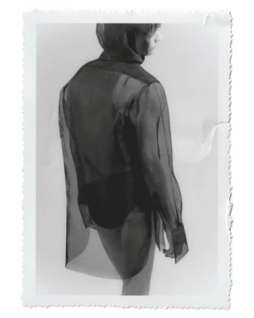 Back view of a slim person wearing a sheer black blouse and dark bottoms, captured in a studio setting under bright, neutral lighting.