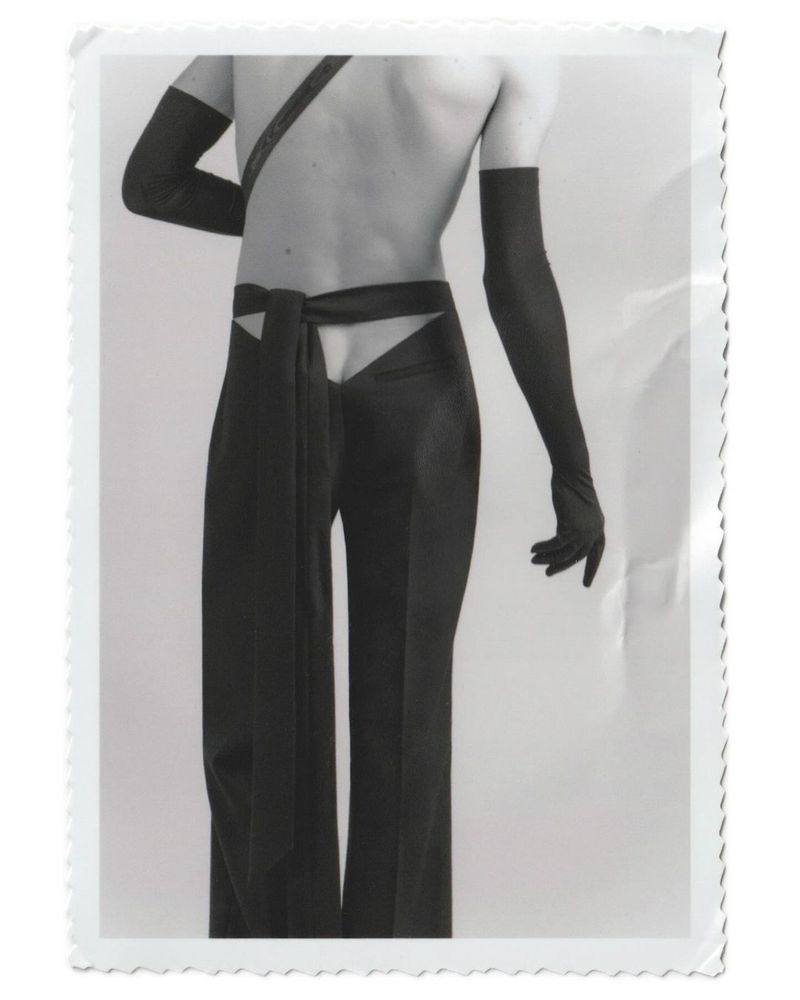 Fashion photograph of a slender adult with bare torso and long black gloves, wearing high-waisted black pants with cut-out detail and sash.