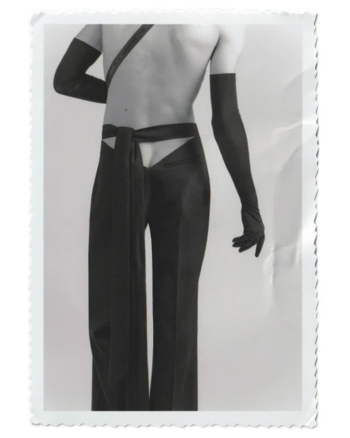 Fashion photograph of a slender adult with bare torso and long black gloves, wearing high-waisted black pants with cut-out detail and sash.