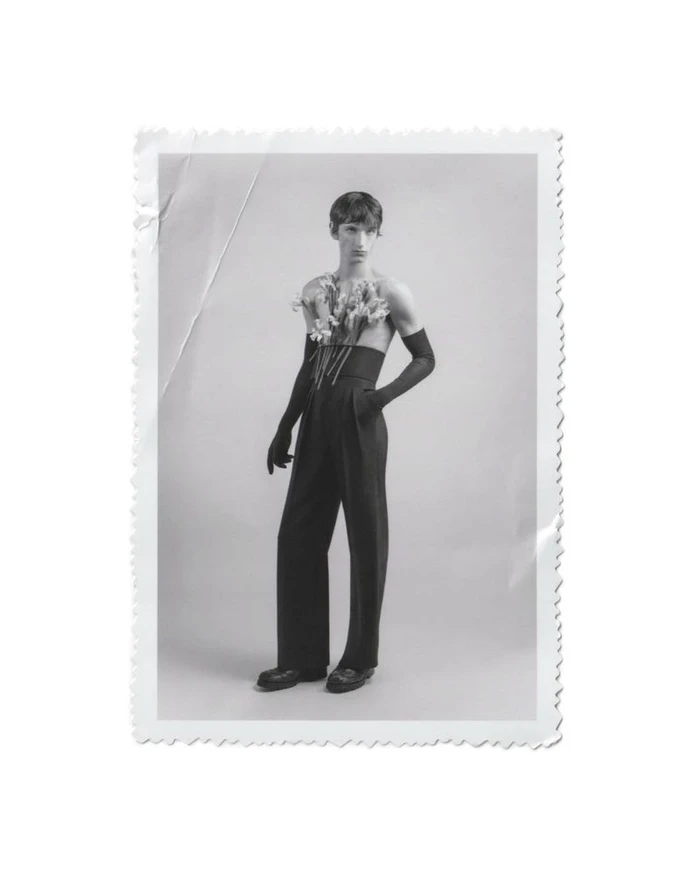 Black and white portrait of a male model holding flowers tucked into high waisted trousers with long gloves against a seamless background.