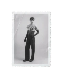 Black and white portrait of a male model holding flowers tucked into high waisted trousers with long gloves against a seamless background.