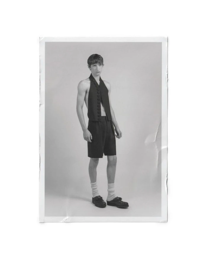Monochrome portrait of a model standing in a black halter-neck vest and tailored shorts against a studio backdrop.
