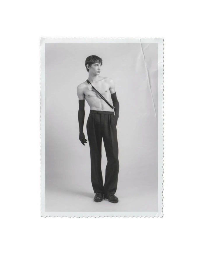 Monochrome portrait of a male model wearing black trousers and long black gloves against a plain studio background.