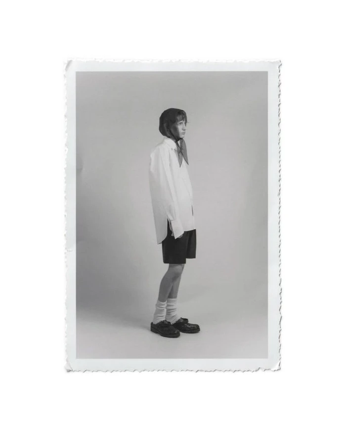 Monochrome portrait of a model standing in profile wearing an oversized white shirt and dark shorts against a seamless studio background.