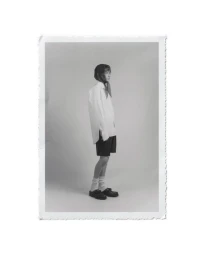 Monochrome portrait of a model standing in profile wearing an oversized white shirt and dark shorts against a seamless studio background.