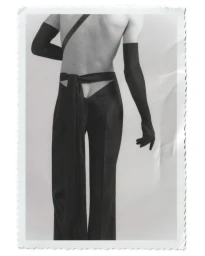 Monochrome rear-view portrait of a model wearing black trousers and opera gloves against a white studio background.