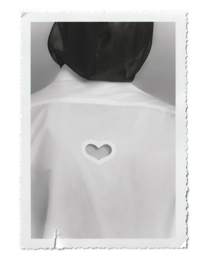 Rear view of a model in a white collared shirt featuring a heart-shaped cutout on the back, wearing a sheer black head covering.