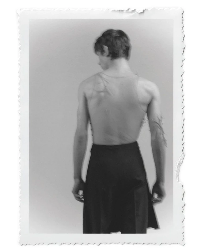Monochrome portrait of a male model seen from behind wearing a tank top and dark skirt against a plain studio background.