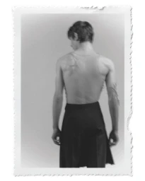 Monochrome portrait of a male model seen from behind wearing a tank top and dark skirt against a plain studio background.