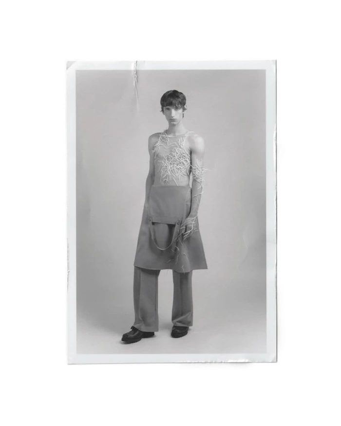 Full-length portrait of a male model wearing a textured top and skirted trousers against a plain studio background.