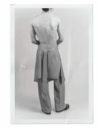 Black and white portrait of a male model from behind wearing trousers with hanging fabric panel and sleeveless top