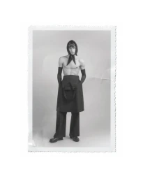 Black and white full-length studio portrait of a shirtless male model wearing a headscarf, gloves, apron skirt, and wide-leg trousers.