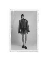 Black and white portrait of a male model wearing a sheer hooded shirt and dark underwear against a seamless white studio backdrop.