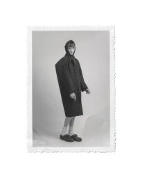 Model Tristan Gibert standing in an oversized dark buttoned coat and headpiece on a light studio background with a deckled edge.
