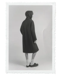 Model Tristan Gibert standing in a dark hooded coat, white socks, and dark shoes against a plain studio backdrop.