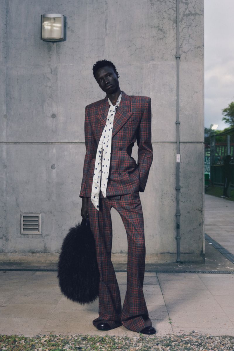 An adult female fashion model poses in a patterned suit, holding a black furry purse outdoors with a concrete wall background.