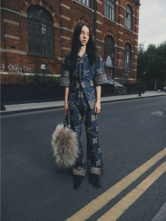 Fashion shot of woman on asphalt street in patterned blue outfit and furry bag, standing near a brick building with muted lighting.