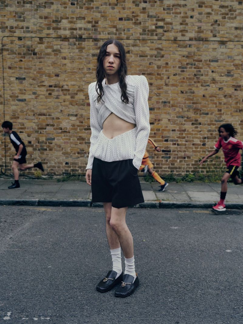 Fashionable person poses on urban street with children playing sports in the background, wearing a cropped white shirt and black shorts.