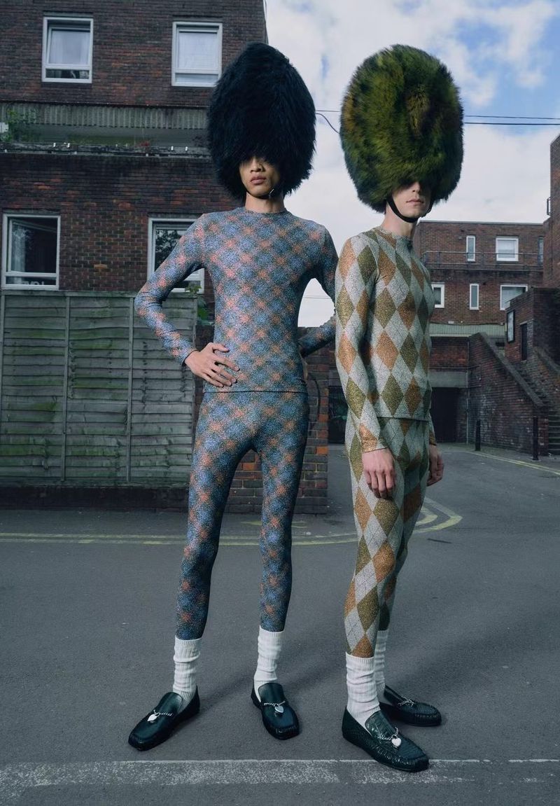Two figures with oversized hats, wearing argyle pattern suits and black loafers, stand on a street in front of a brick apartment building.