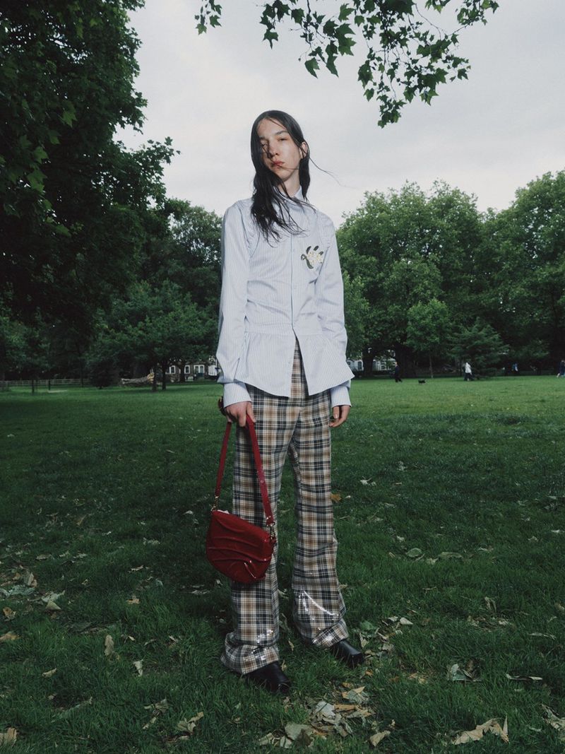 A young woman in a button-down shirt and plaid pants stands in a park, holding a red purse, with trees and grass surrounding her.