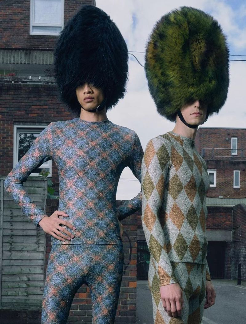 Two people wearing matching diamond-patterned knitwear and large fuzzy hats in front of a brick building on a city street.