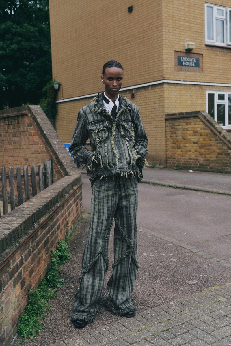 A young, dark-skinned male fashion model posing outdoors wearing a distressed green and black plaid suit in front of a brick building.